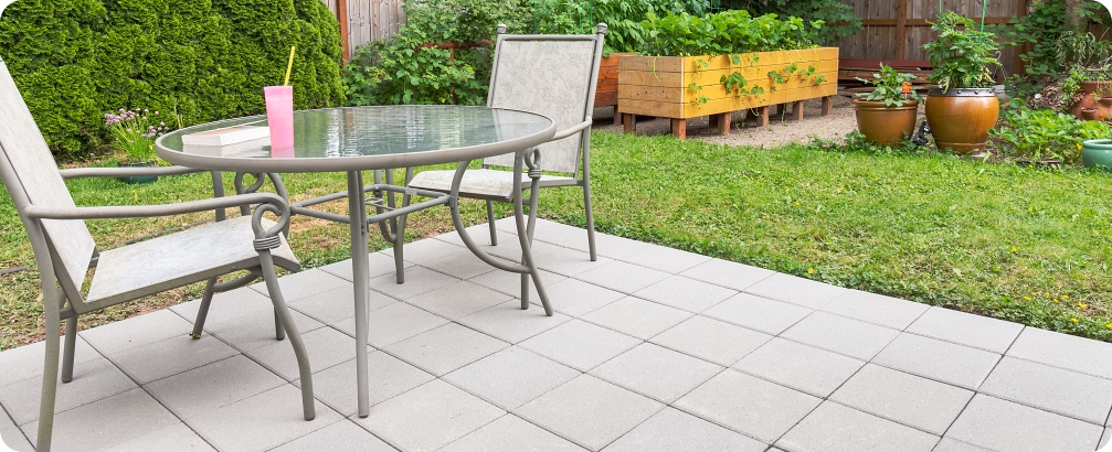 Outdoor patio with a glass-top table and chairs overlooking a landscaped backyard with a green lawn, raised garden beds, and potted plants