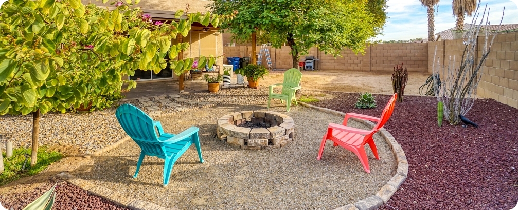 Landscaped backyard with a circular stone fire pit, colorful outdoor chairs, gravel ground cover, mature trees, and desert-style plants