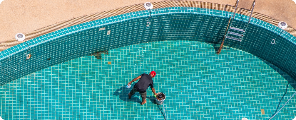 Pool maintenance worker cleaning the bottom of an empty swimming pool with turquoise tile flooring.